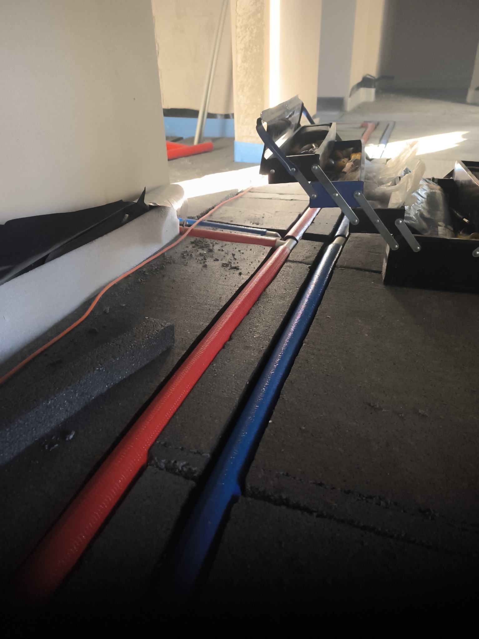 Czerwona i niebieska rura instalacji grzewczej ułożona w bruzdach w podłodze, obok rozłożona skrzynka z narzędziami.