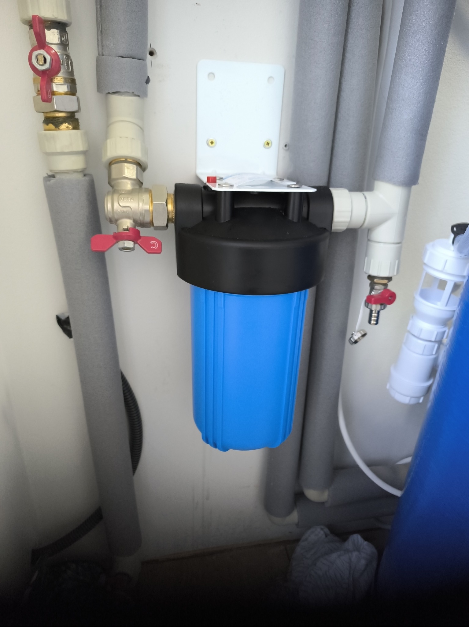 Filtr wody z niebieskim korpusem, zawieszony na białej ścianie, otoczony rurami z szarą izolacją i zaworami z czerwonymi uchwytami, element instalacji wodnej.