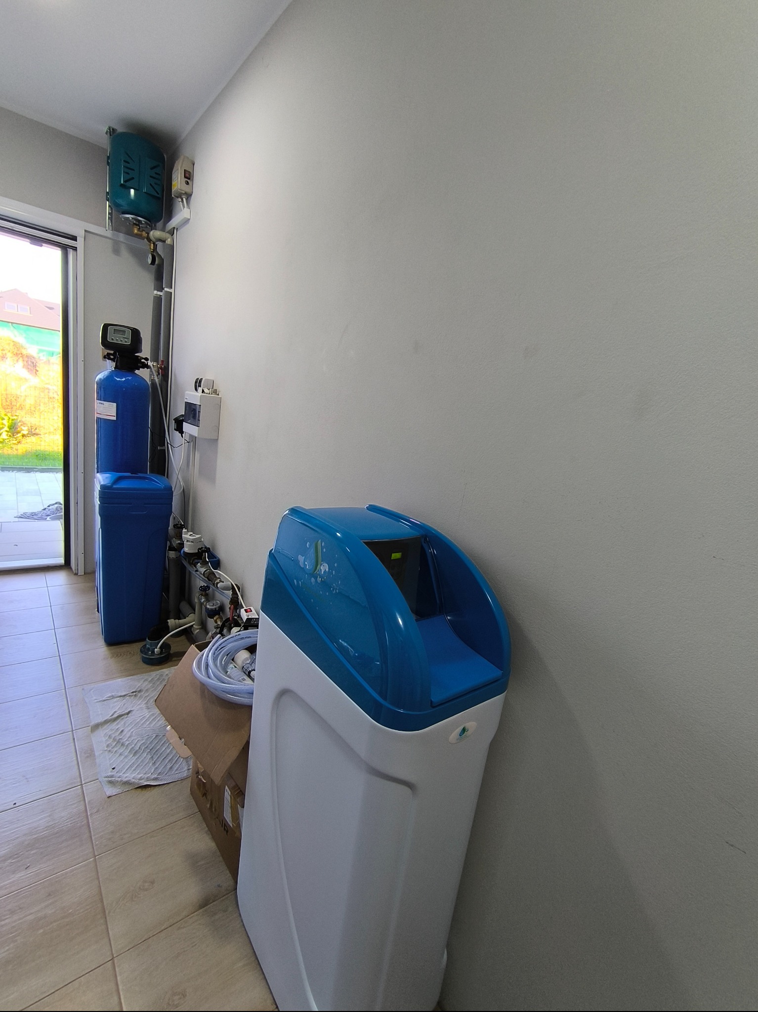 Biało-niebieski zmiękczacz wody stoi w rogu pomieszczenia, widoczne również elementy instalacji wodnej, rury i zawory, karton na podłodze.