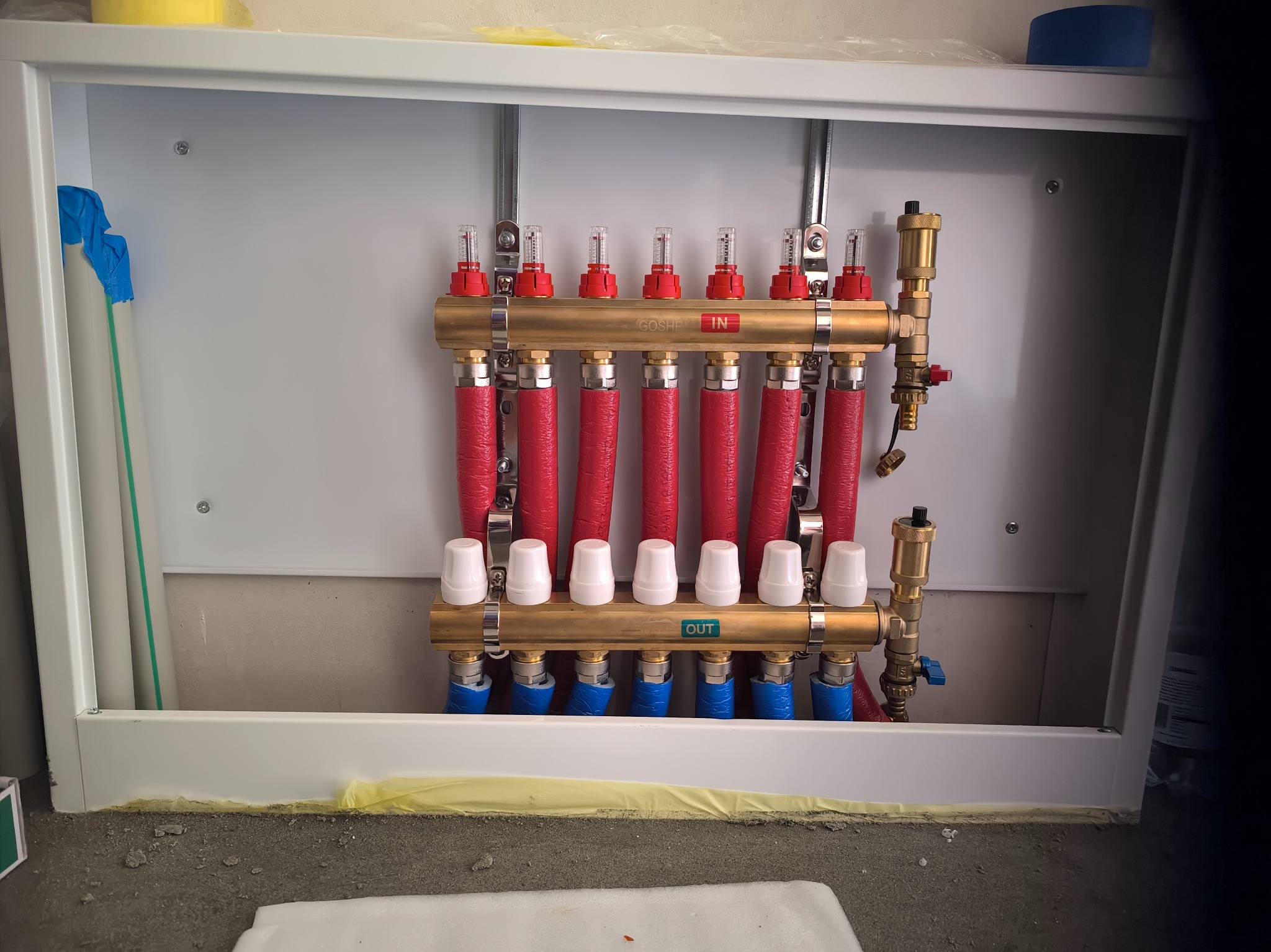 Rozdzielacz ogrzewania podłogowego w białej szafce, z widocznymi rurami czerwonymi (wejście) i niebieskimi (wyjście), oraz białymi termostatami, zamontowany na szarej ścianie z surowego betonu.