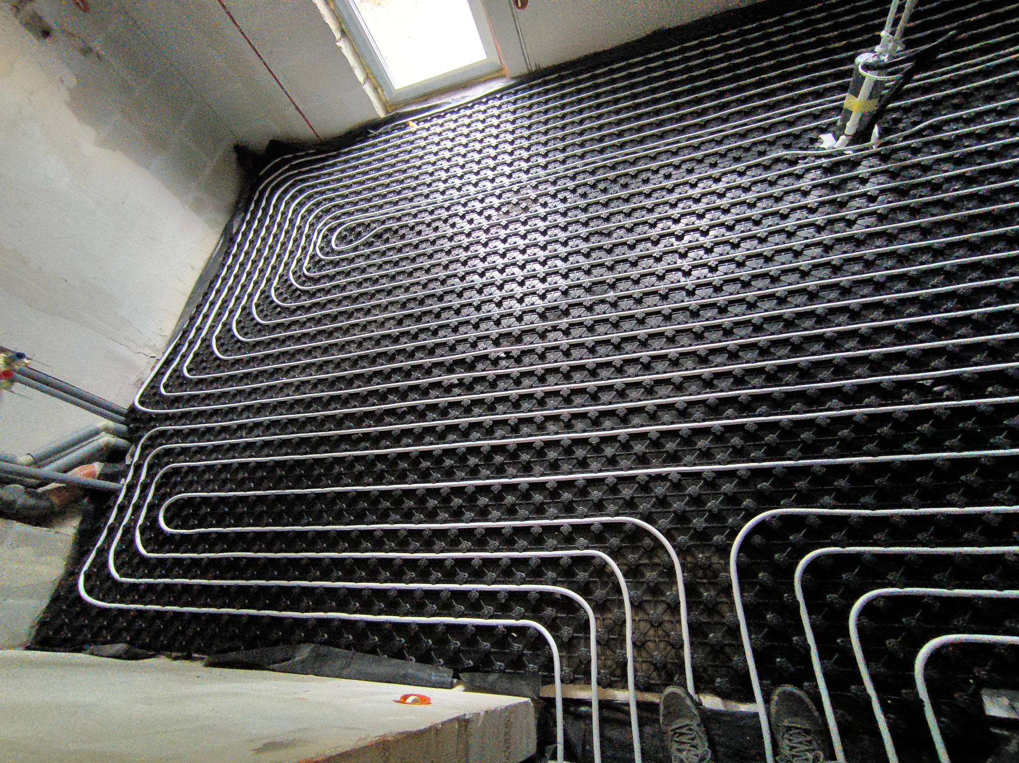 Białe rury ogrzewania podłogowego ułożone spiralnie na czarnej macie izolacyjnej w surowym wnętrzu z widocznym oknem dachowym i fragmentem butów