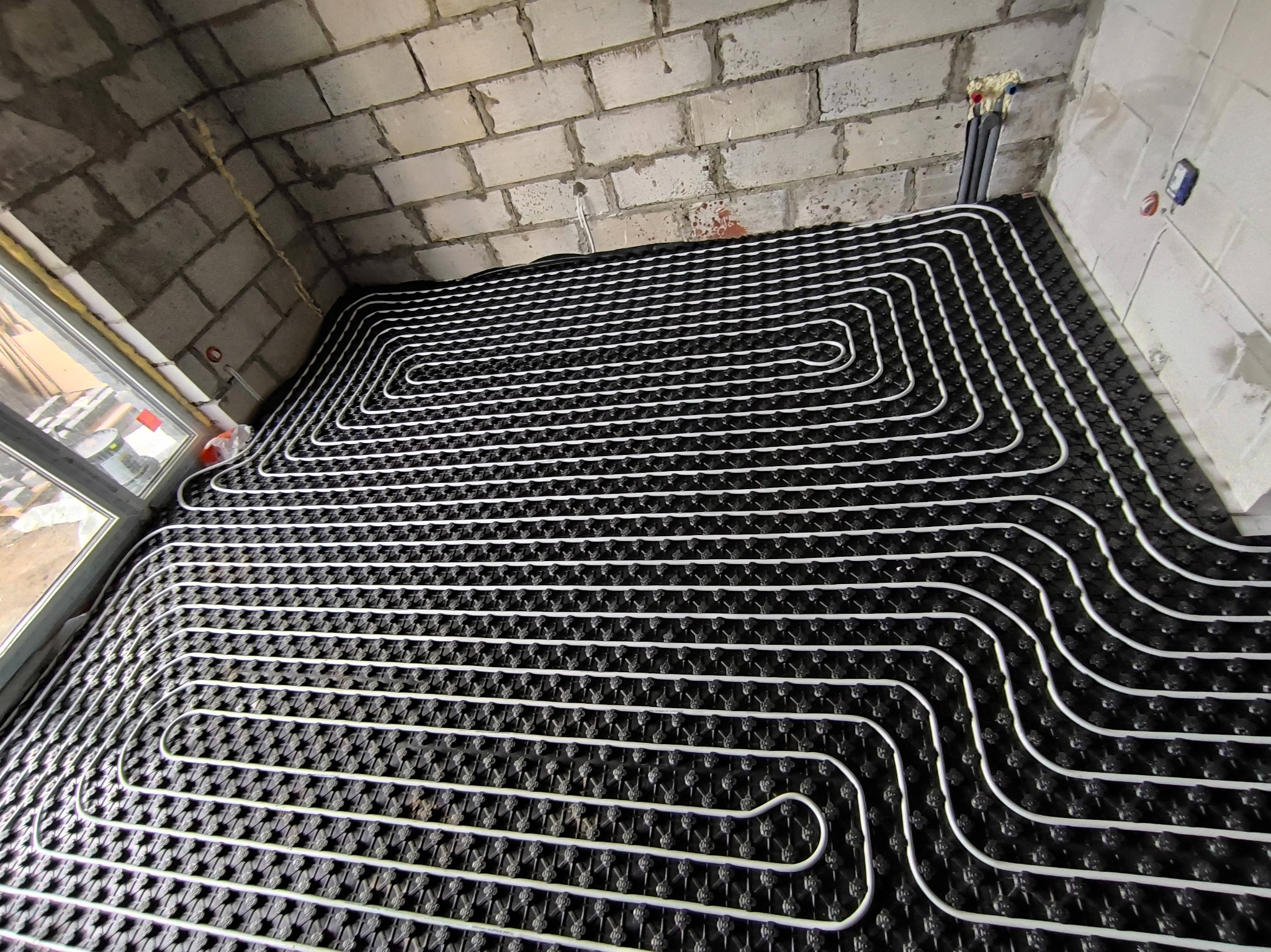 Białe rury grzewcze ułożone w regularnych pętlach na czarnej macie izolacyjnej, widoczne surowe ściany z cegieł i instalacja wodna w tle.