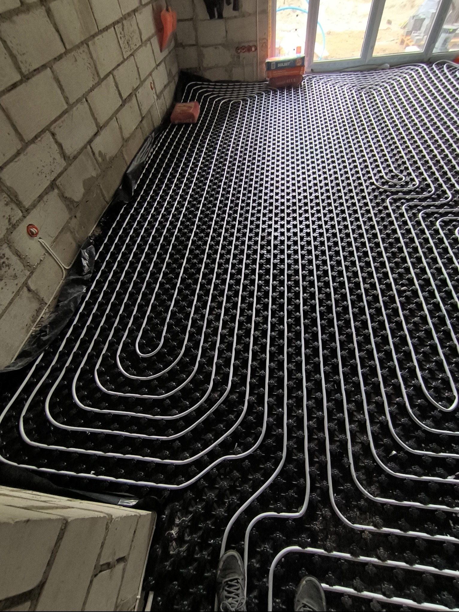 Białe rury ogrzewania podłogowego ułożone na czarnej macie izolacyjnej w surowym pomieszczeniu z ceglanymi ścianami, widoczne stopy w butach w dolnym rogu.