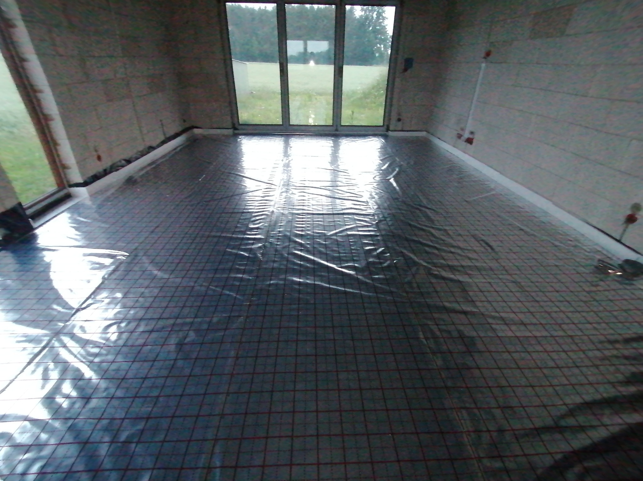 Przygotowanie do montażu ogrzewania podłogowego: rozłożona mata grzewcza z folią ochronną w surowym wnętrzu z widocznymi przyłączami na ścianie.