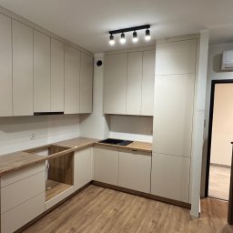 WYMIAROWE STUDIO MEBLI MICHAŁ ZALEWSKI - Wnętrze kuchni z jasnymi, minimalistycznymi meblami. Część blatu roboczego z wyciętym otworem na zlew, widoczne niedoskonałości montażu folii ochronnej na blacie. Na suficie czarna lampa z czterema...