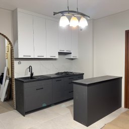 WYMIAROWE STUDIO MEBLI MICHAŁ ZALEWSKI - Nowoczesna kuchnia z białymi szafkami górnymi i grafitowymi dolnymi, czarnym blatem i armaturą. Widoczny fragment wyspy kuchennej, płyta gazowa i okap. W tle łukowate przejście do innego...