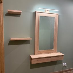 WYMIAROWE STUDIO MEBLI MICHAŁ ZALEWSKI - Drewniana toaletka z lustrem i oświetleniem LED, uzupełniona o dwie półki na ścianie w kolorze oliwkowym, z widocznym gniazdkiem elektrycznym w dolnym rogu.