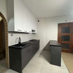 WYMIAROWE STUDIO MEBLI MICHAŁ ZALEWSKI - Nowoczesna kuchnia z białymi szafkami wiszącymi i ciemnoszarymi szafkami dolnymi, czarnym zlewem i blatem, widoczny fragment baru i drzwi z krzyżem, wykończenie ceglane łuku.