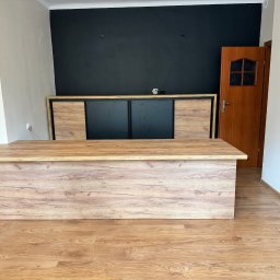 WYMIAROWE STUDIO MEBLI MICHAŁ ZALEWSKI - Biurko z drewnopodobnym blatem i frontem, z szafką za nim na tle czarnej ściany, w pokoju z panelami podłogowymi.