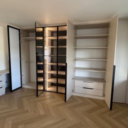 WYMIAROWE STUDIO MEBLI MICHAŁ ZALEWSKI - Jasna szafa z oświetleniem LED i czarnymi ramami, otwarte drzwi ukazują półki i szuflady. Parkiet w jodełkę na podłodze. Widoczna komoda z szufladami.