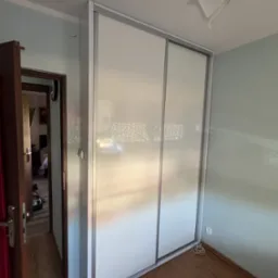 Biała szafa wnękowa z przesuwnymi drzwiami w pokoju z drewnianą podłogą. Widoczny fragment ściany w kolorze jasnego błękitu i lampa sufitowa.