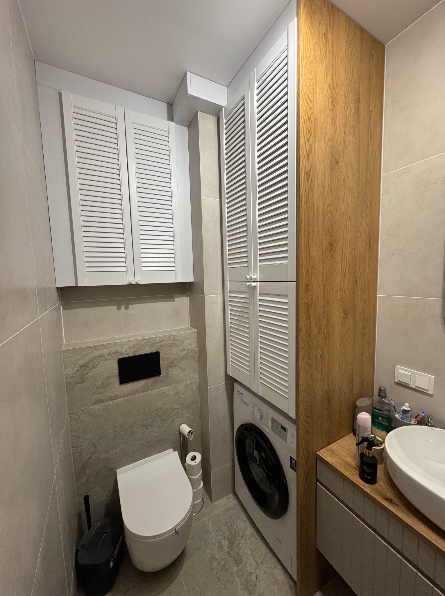 Nowoczesna łazienka z zabudową meblową: szafki z lamelowymi frontami nad pralką i toaletą, umywalka nablatowa na drewnianym blacie, szara glazura, minimalistyczny design.