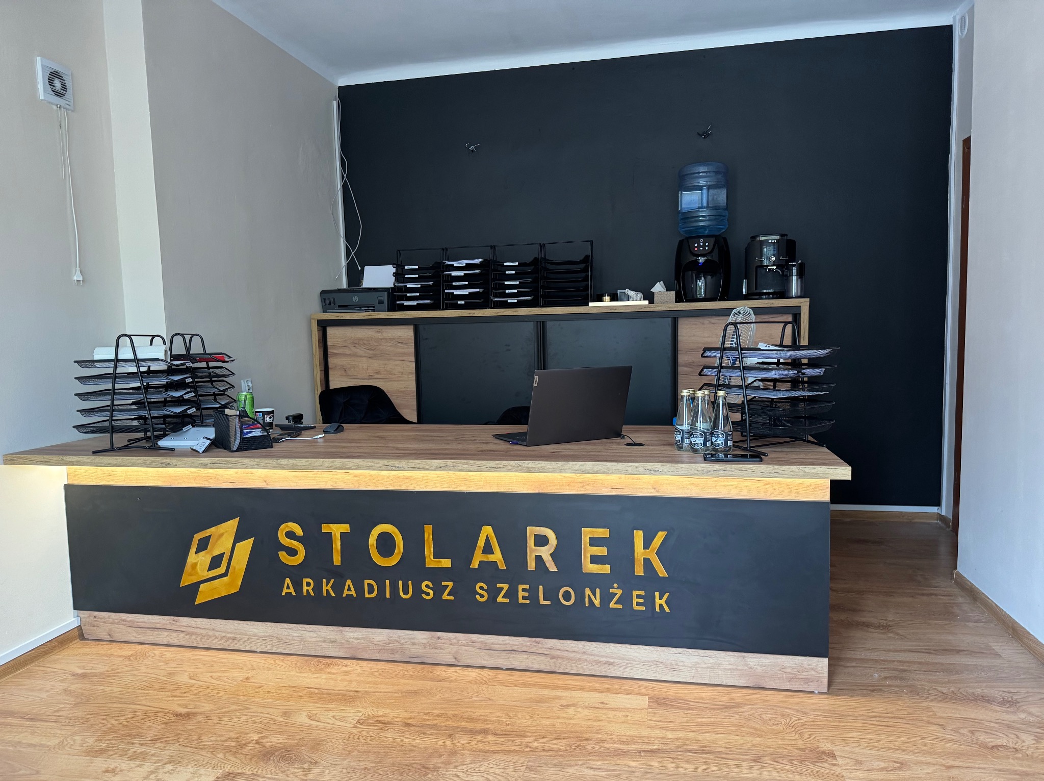 Biuro z recepcją, na której widnieje napis 'Stolarek Arkadiusz Szelążek' złotymi literami na czarnym tle, laptop, podajniki na dokumenty, dystrybutor wody i ekspres do kawy.