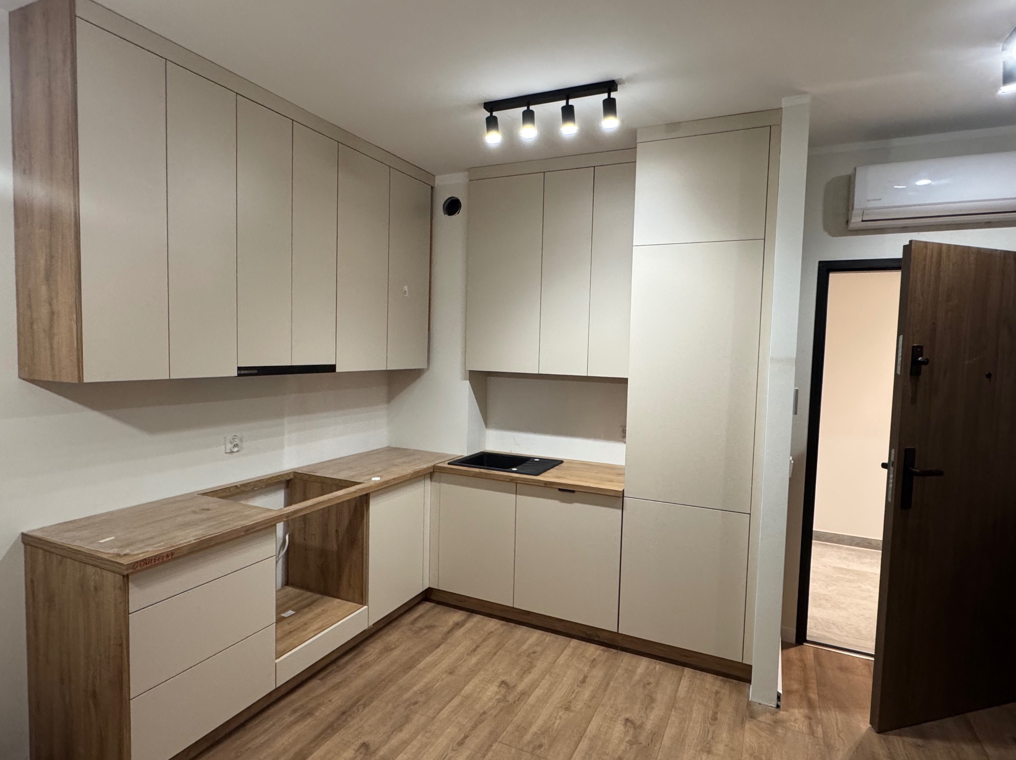 Wnętrze kuchni z jasnymi, minimalistycznymi meblami. Część blatu roboczego z wyciętym otworem na zlew, widoczne niedoskonałości montażu folii ochronnej na blacie. Na suficie czarna lampa z czterema...