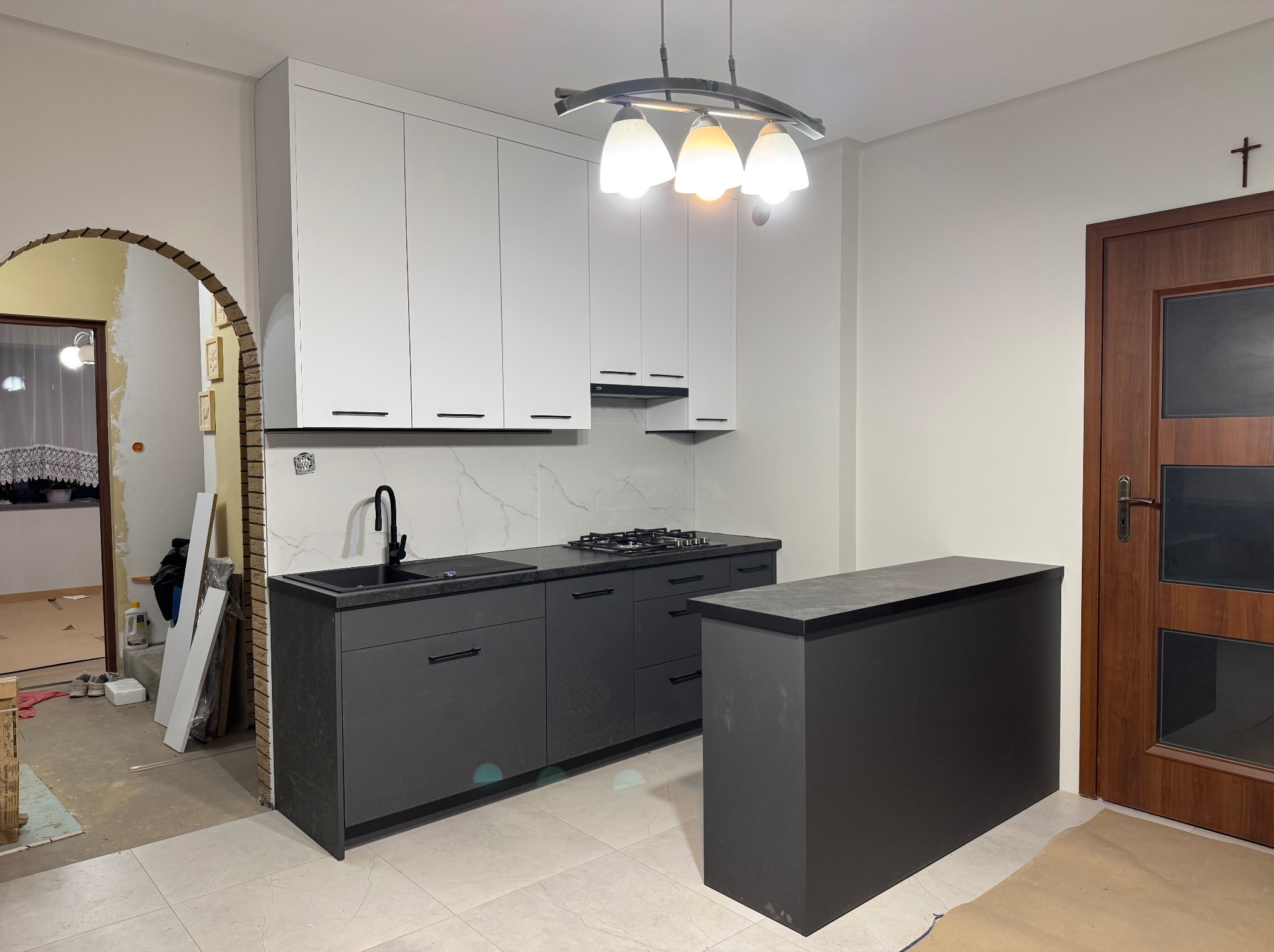 Nowoczesna kuchnia z białymi szafkami górnymi i grafitowymi dolnymi, czarnym blatem i armaturą. Widoczny fragment wyspy kuchennej, płyta gazowa i okap. W tle łukowate przejście do innego...