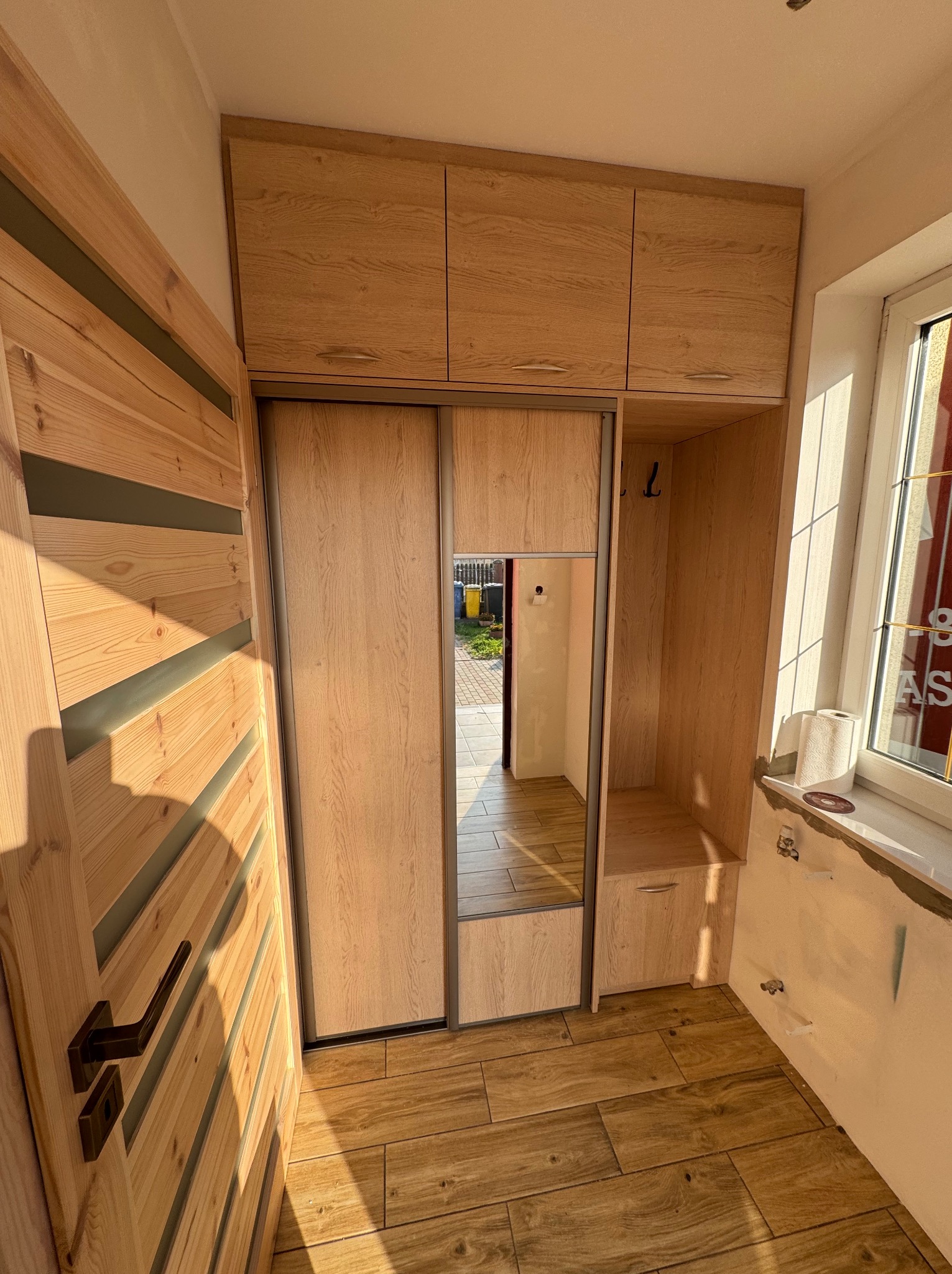 Wnęka z drewnianą szafą z przesuwnymi drzwiami z lustrem, nad nią szafki wiszące, obok wnęka z wieszakami i siedziskiem, drewniane panele na podłodze, widoczna drewniana drzwi z poziomymi...