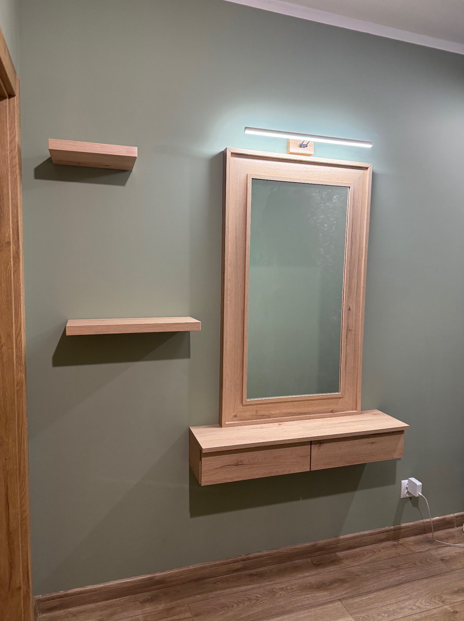 Drewniana toaletka z lustrem i oświetleniem LED, uzupełniona o dwie półki na ścianie w kolorze oliwkowym, z widocznym gniazdkiem elektrycznym w dolnym rogu.