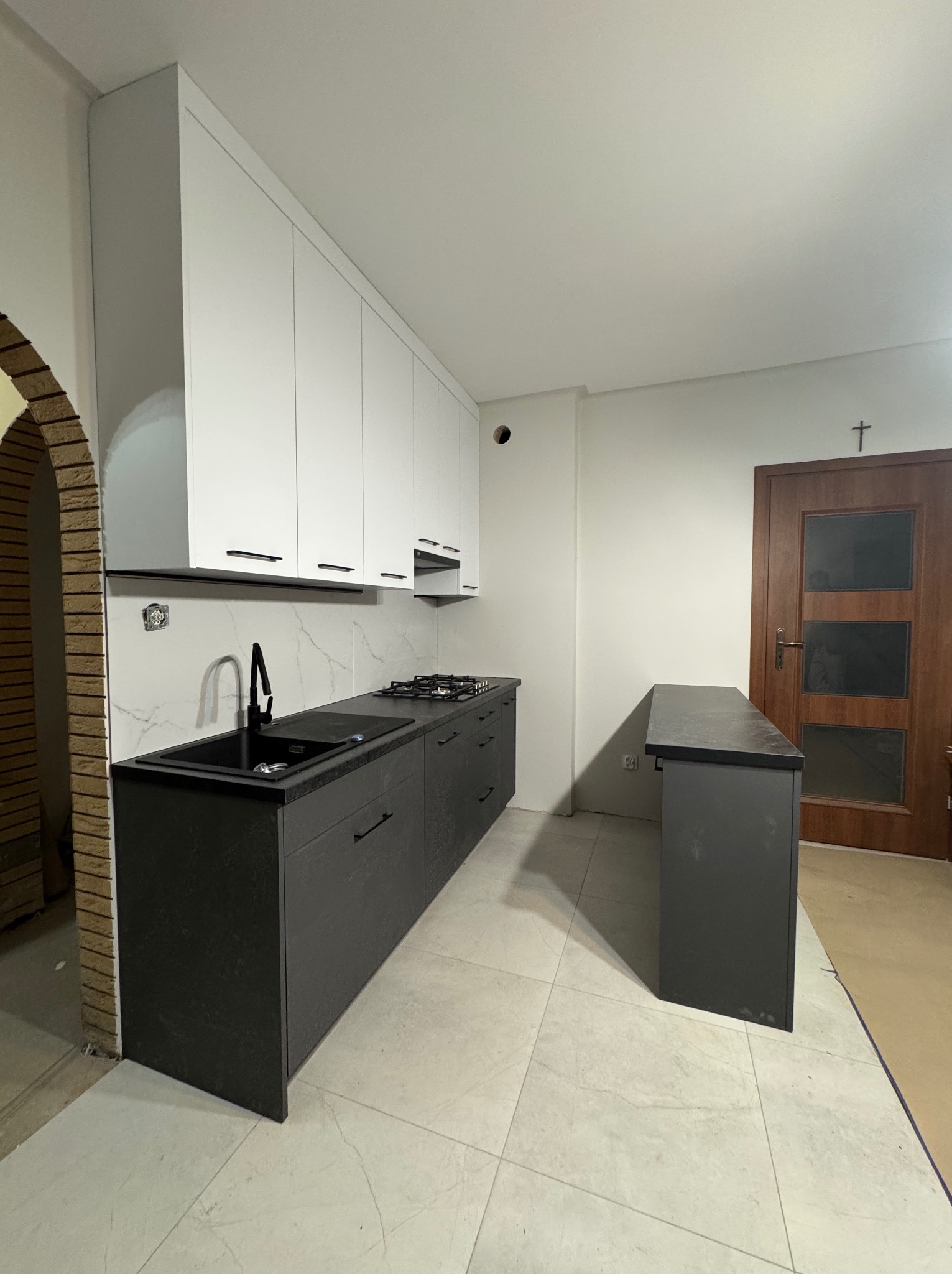 Nowoczesna kuchnia z białymi szafkami wiszącymi i ciemnoszarymi szafkami dolnymi, czarnym zlewem i blatem, widoczny fragment baru i drzwi z krzyżem, wykończenie ceglane łuku.