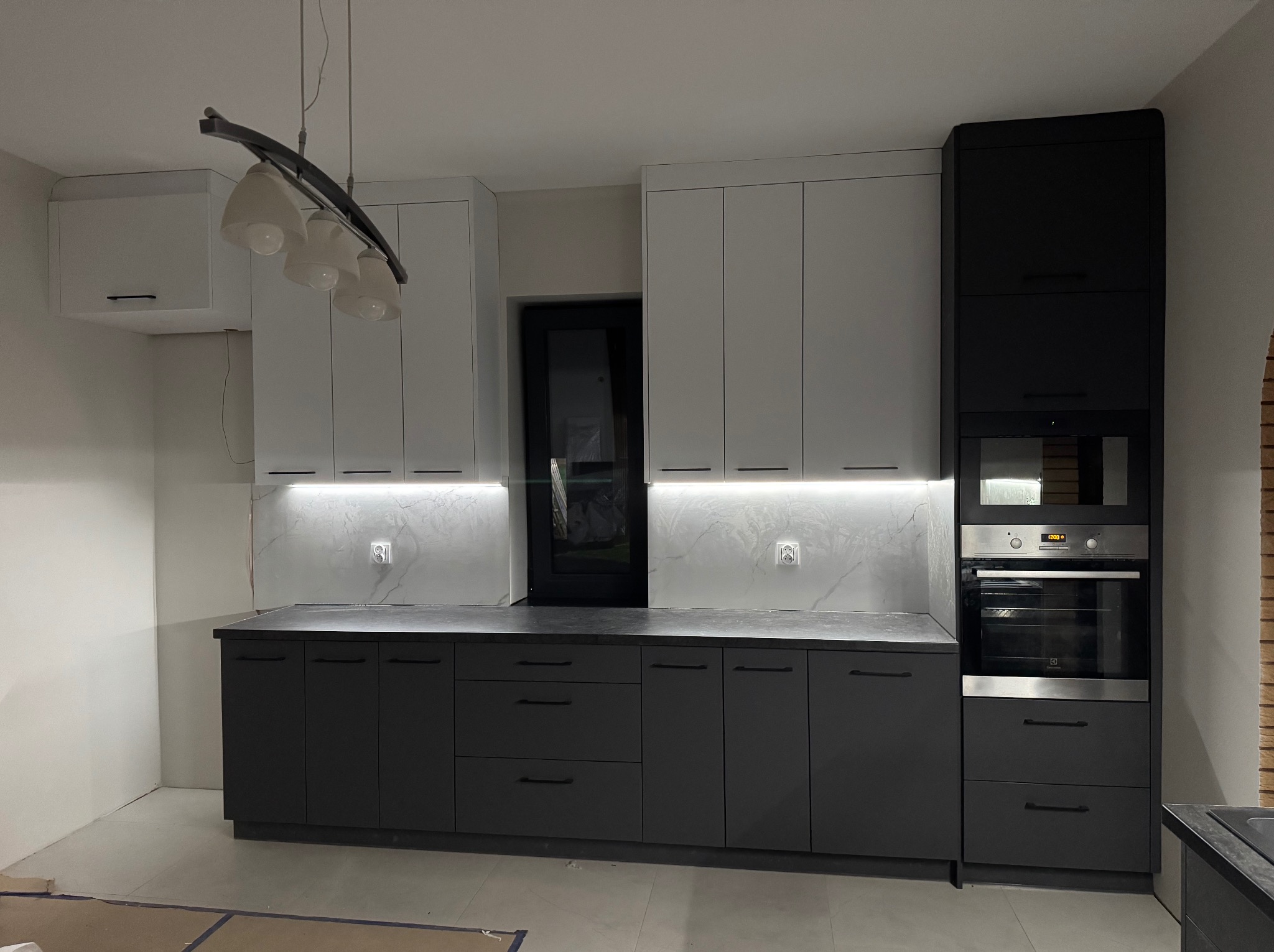 Nowoczesna kuchnia z białymi górnymi szafkami i grafitowymi dolnymi, oświetlenie LED pod szafkami, czarne okno w tle, lampa wisząca z kilkoma białymi kloszami.