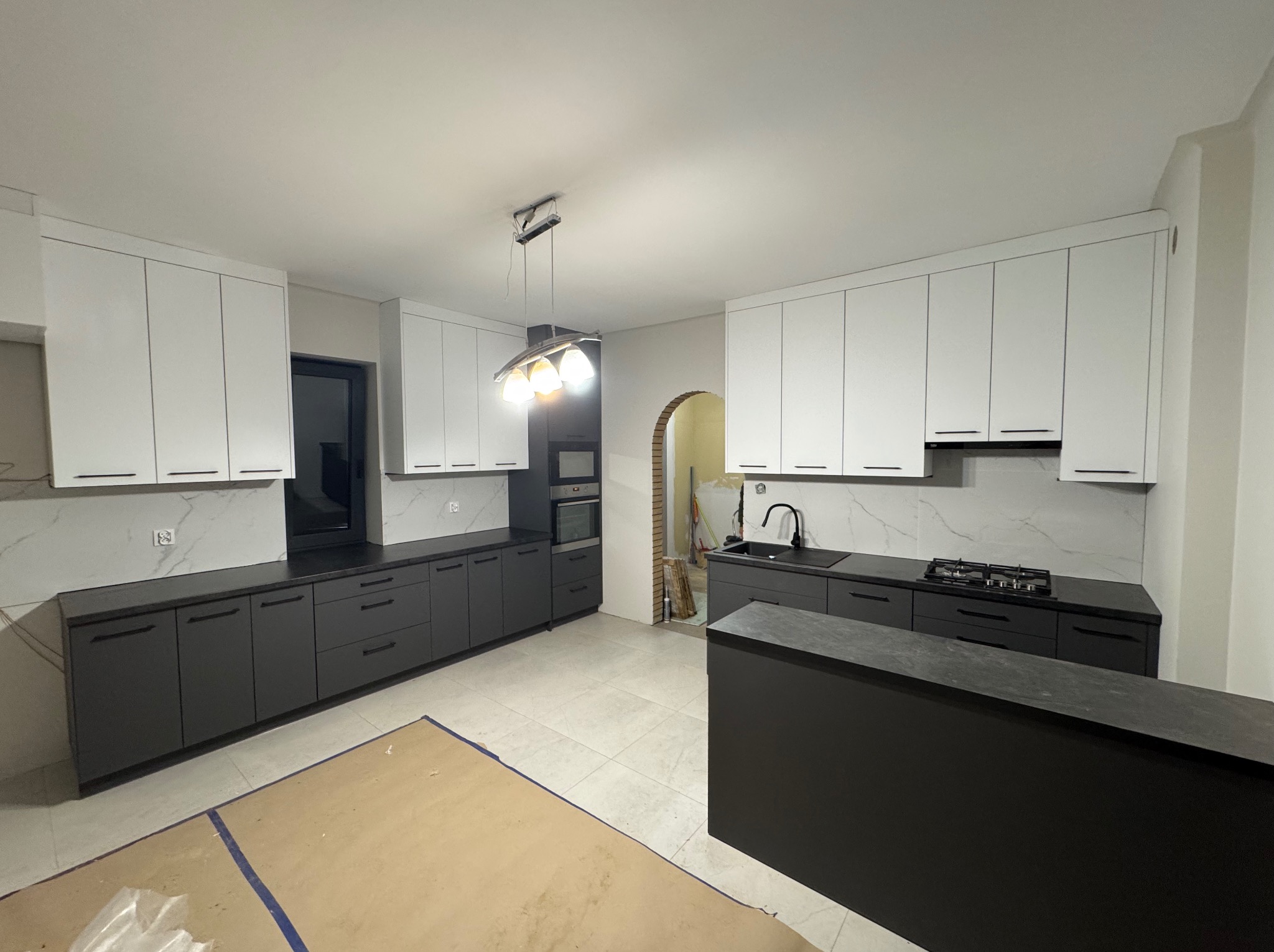 Nowoczesna kuchnia z białymi szafkami górnymi i grafitowymi dolnymi, czarnym blatem oraz marmurowym backsplashiem, widok na łuk w ścianie prowadzący do innego pomieszczenia, podłoga częściowo...