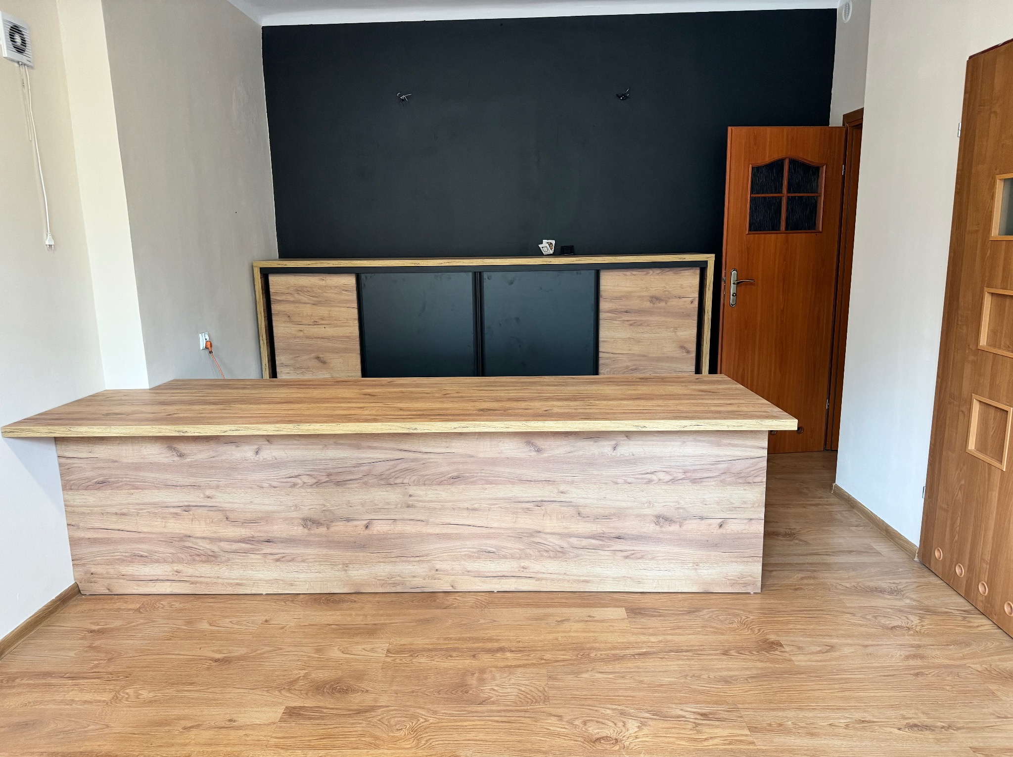 Biurko z drewnopodobnym blatem i frontem, z szafką za nim na tle czarnej ściany, w pokoju z panelami podłogowymi.