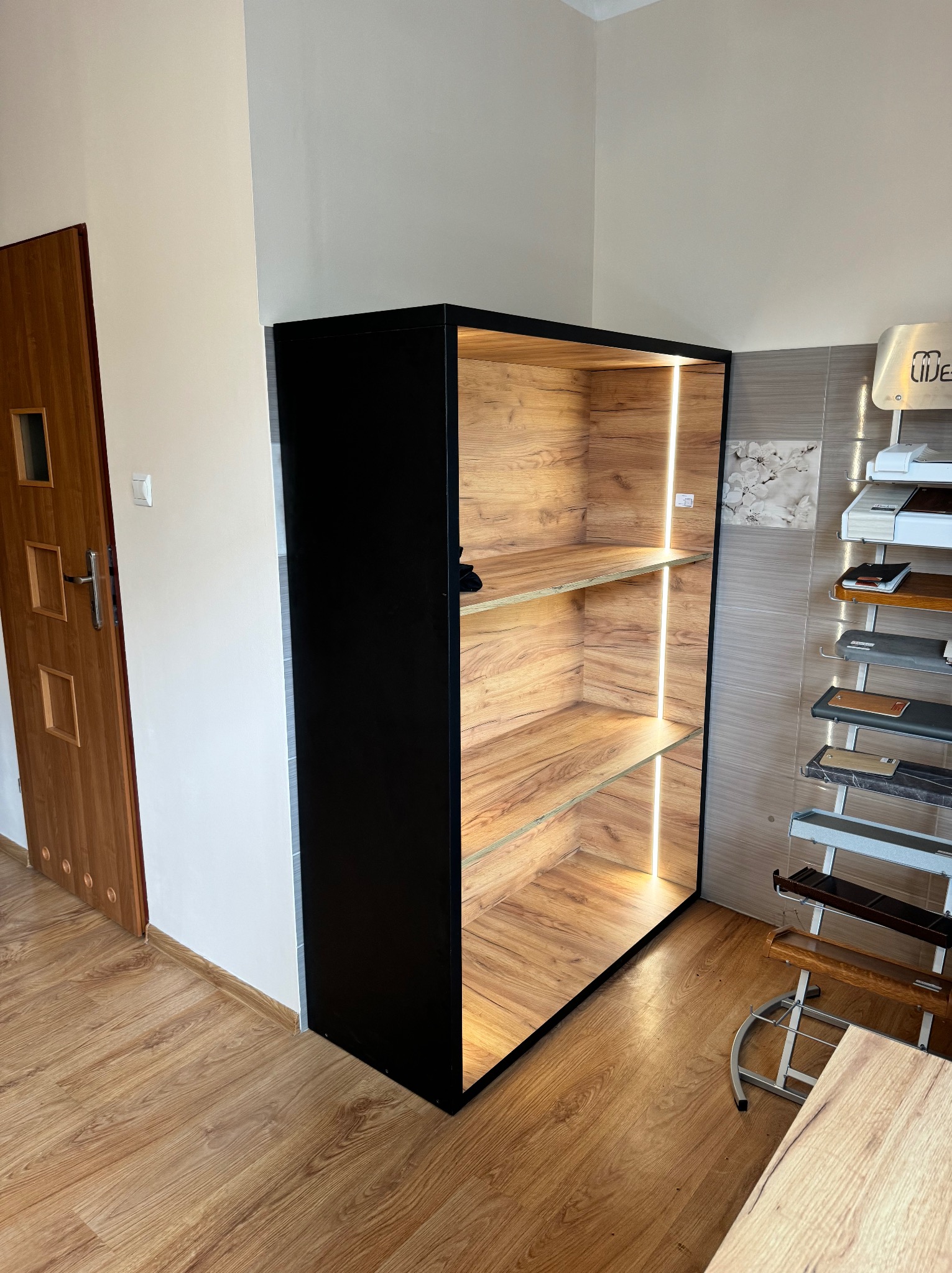 Czarna szafa wnękowa z oświetleniem LED, wykończona drewnopodobną okleiną, stojąca na drewnianej podłodze, obok ekspozytor z próbkami materiałów wykończeniowych i brązowe drzwi.