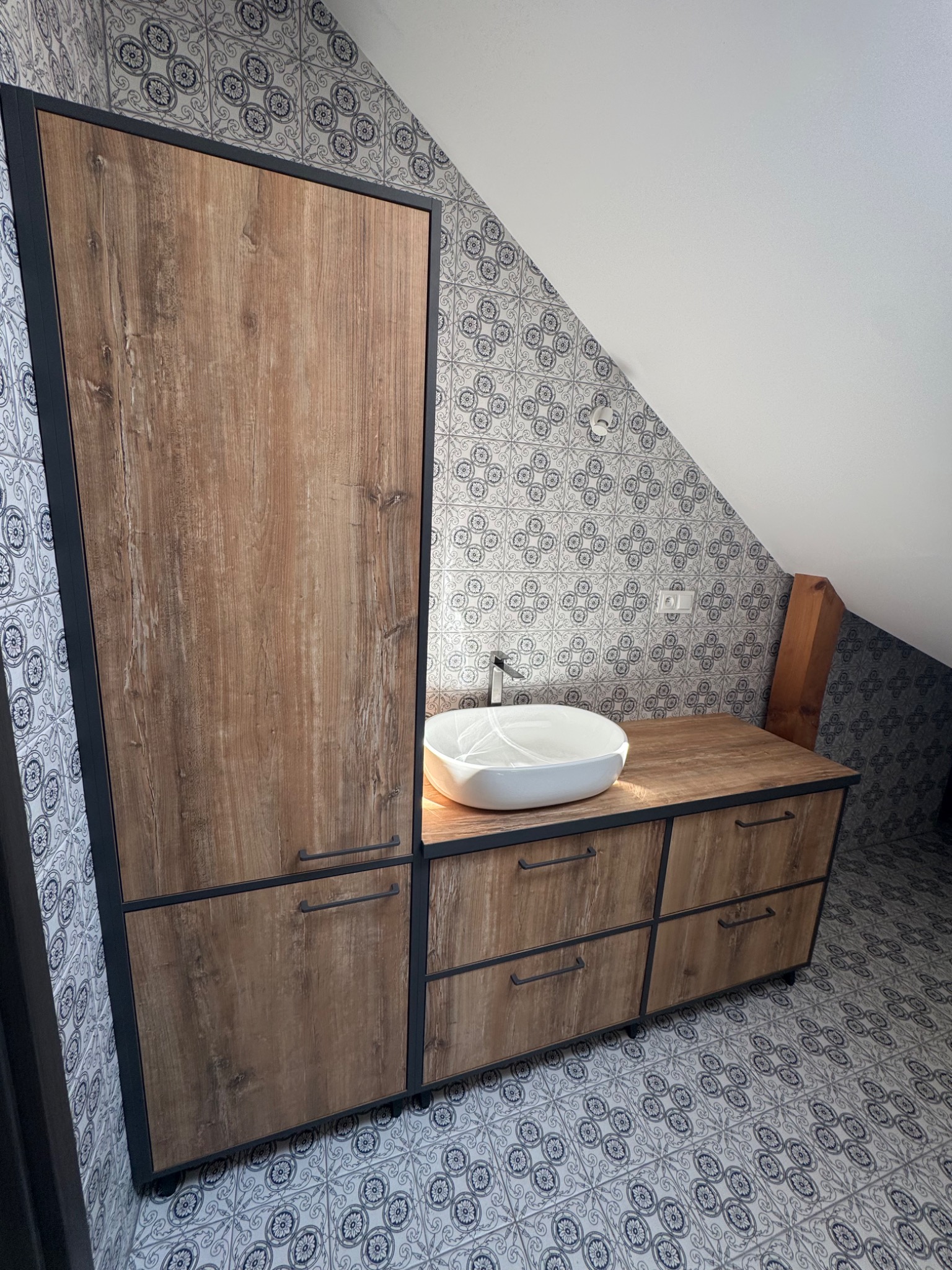 Nowoczesna zabudowa łazienkowa z drewnianymi frontami i czarnymi ramami, umywalka nablatowa, wzorzyste płytki na ścianach i podłodze w łazience na poddaszu.