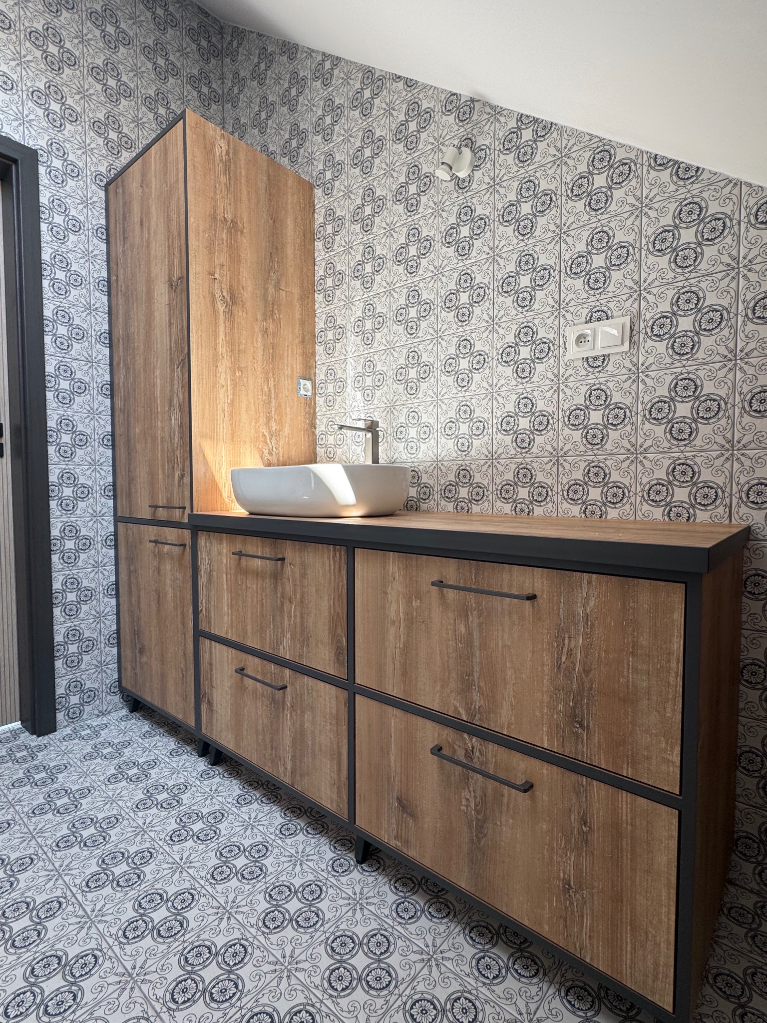 Nowoczesna szafka łazienkowa z umywalką nablatową i drewnianymi frontami na tle wzorzystych płytek. Czarna rama i uchwyty dodają elegancji.