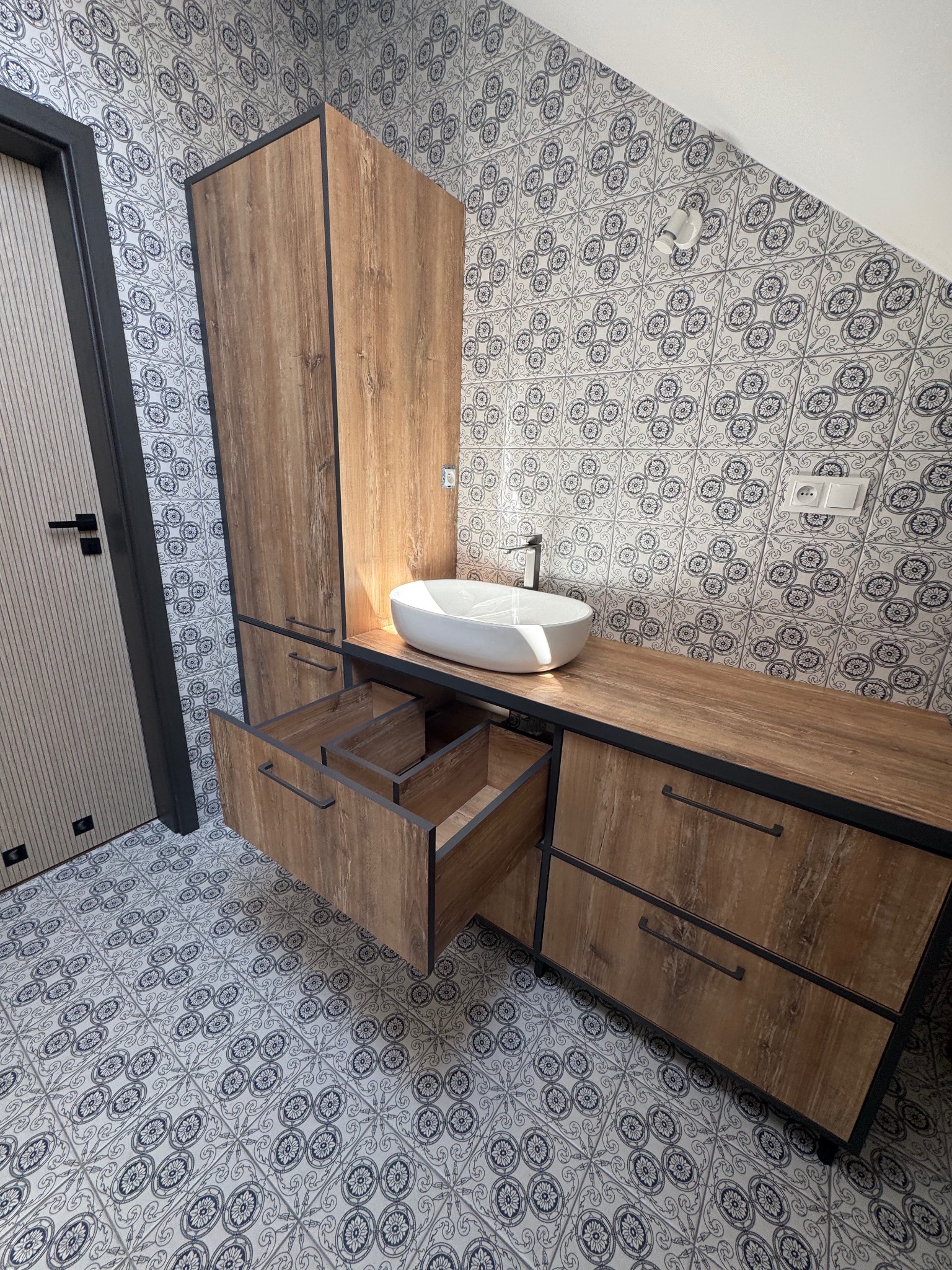 Nowoczesna łazienka z drewnianą szafką pod umywalkę z otwartymi szufladami, czarnymi detalami i wzorzystymi płytkami na ścianach i podłodze.