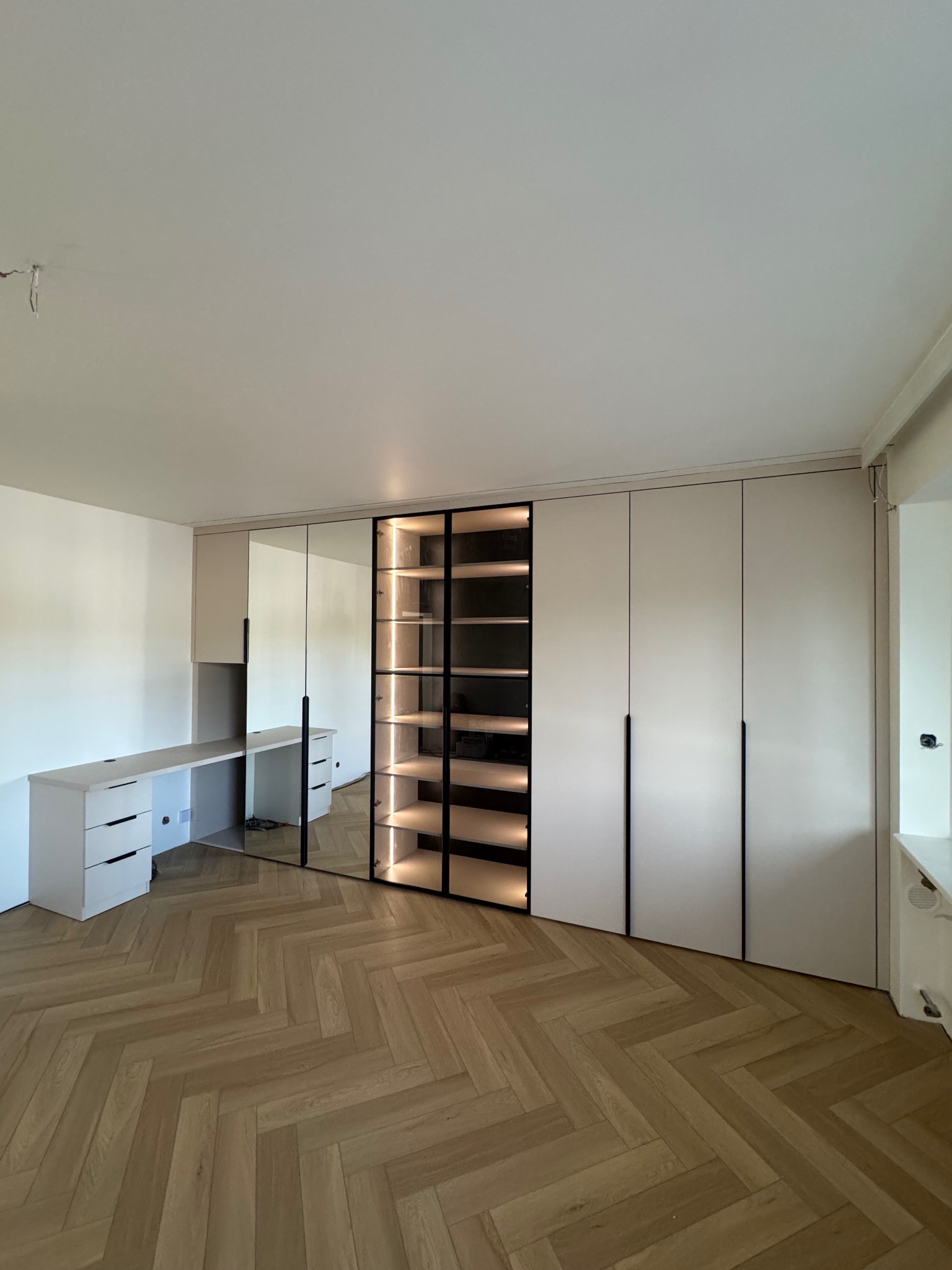 Jasna szafa wnękowa z biurkiem i podświetlaną witryną, parkiet w jodełkę. Minimalistyczny design, czarne uchwyty. Nowoczesne meble na wymiar.