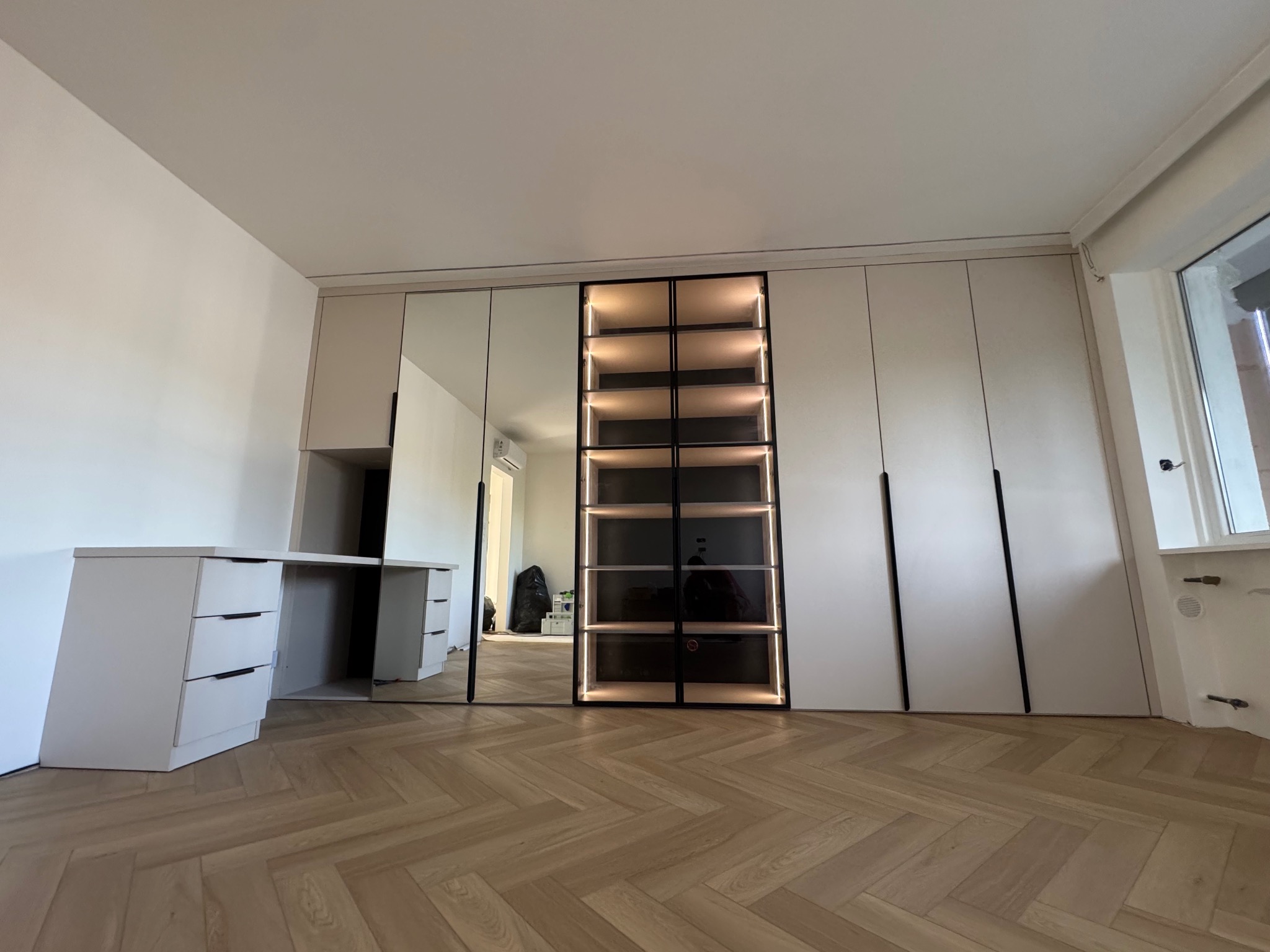 Nowoczesna szafa wnękowa z lustrami i oświetleniem LED, połączona z biurkiem, na parkiecie ułożonym w jodełkę, w jasnym wnętrzu.