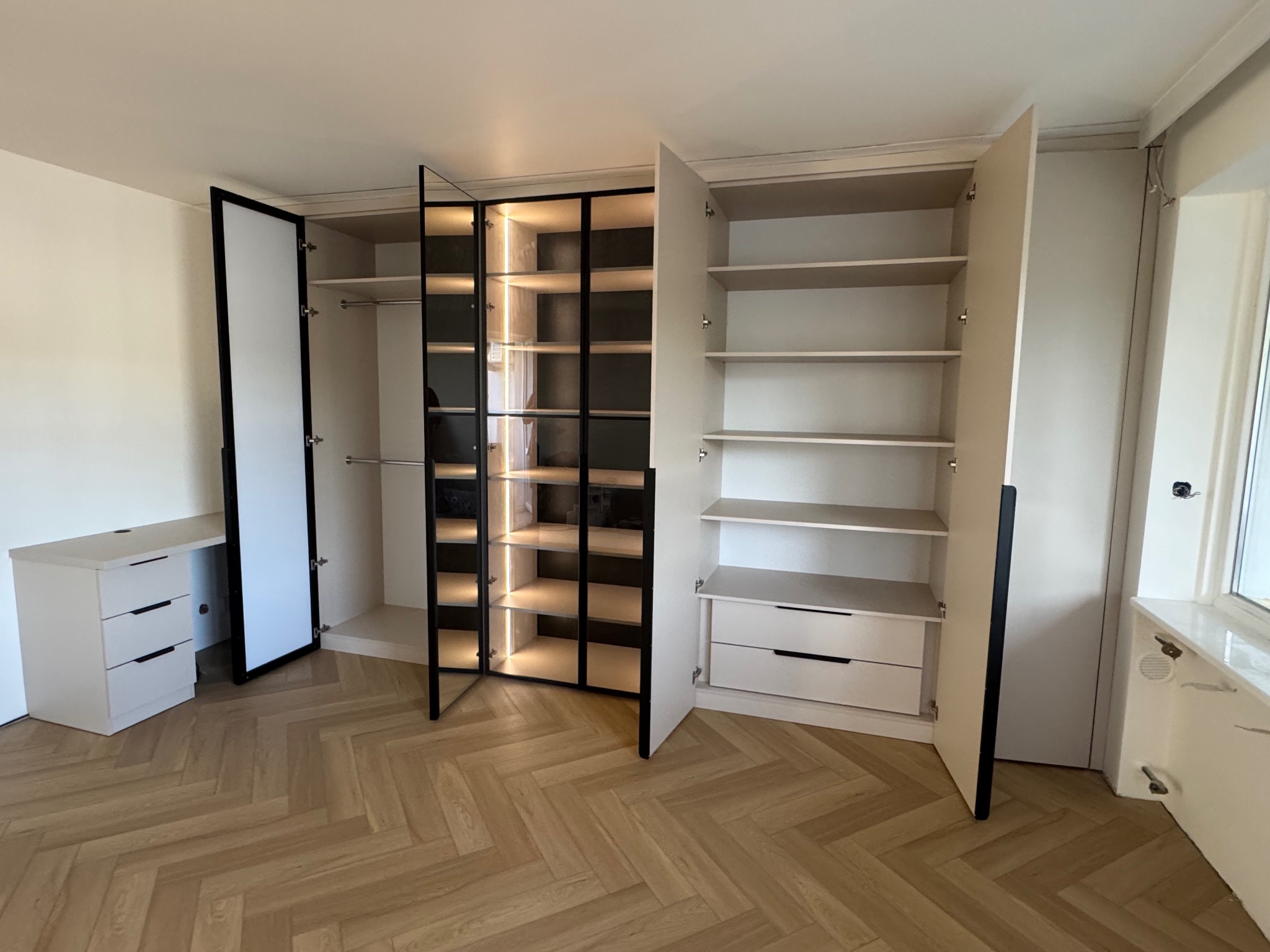 Jasna szafa z oświetleniem LED i czarnymi ramami, otwarte drzwi ukazują półki i szuflady. Parkiet w jodełkę na podłodze. Widoczna komoda z szufladami.