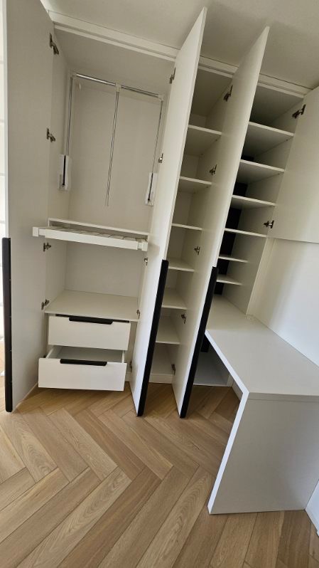 Biała szafa wnękowa z czarnymi akcentami i wysuwanymi szufladami na parkiecie w jodełkę. Otwarta, ukazująca półki i drążki na ubrania.