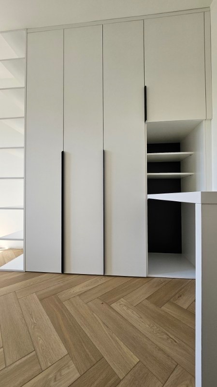 Biała szafa wnękowa z czarnymi uchwytami i otwartą półką z czarnym tłem, na podłodze parkiet ułożony w jodełkę.