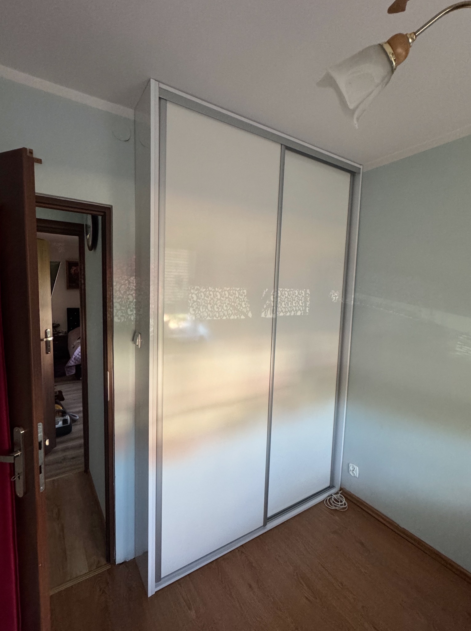 Biała szafa wnękowa z przesuwnymi drzwiami w pokoju z drewnianą podłogą. Widoczny fragment ściany w kolorze jasnego błękitu i lampa sufitowa.