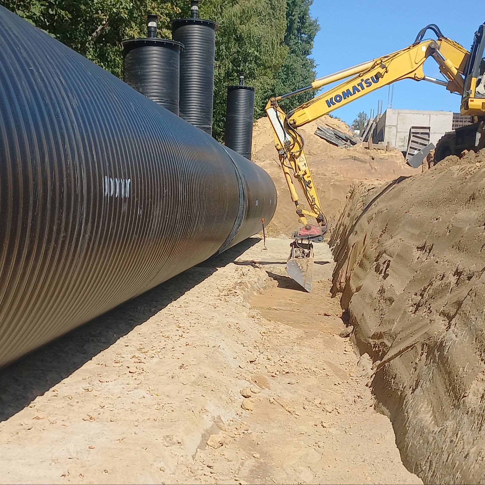 Żółta koparka Komatsu przygotowuje wykop pod dużą, czarną, karbowaną rurę kanalizacyjną; w tle widoczne pionowe kominy wentylacyjne i konstrukcja budynku w trakcie budowy.