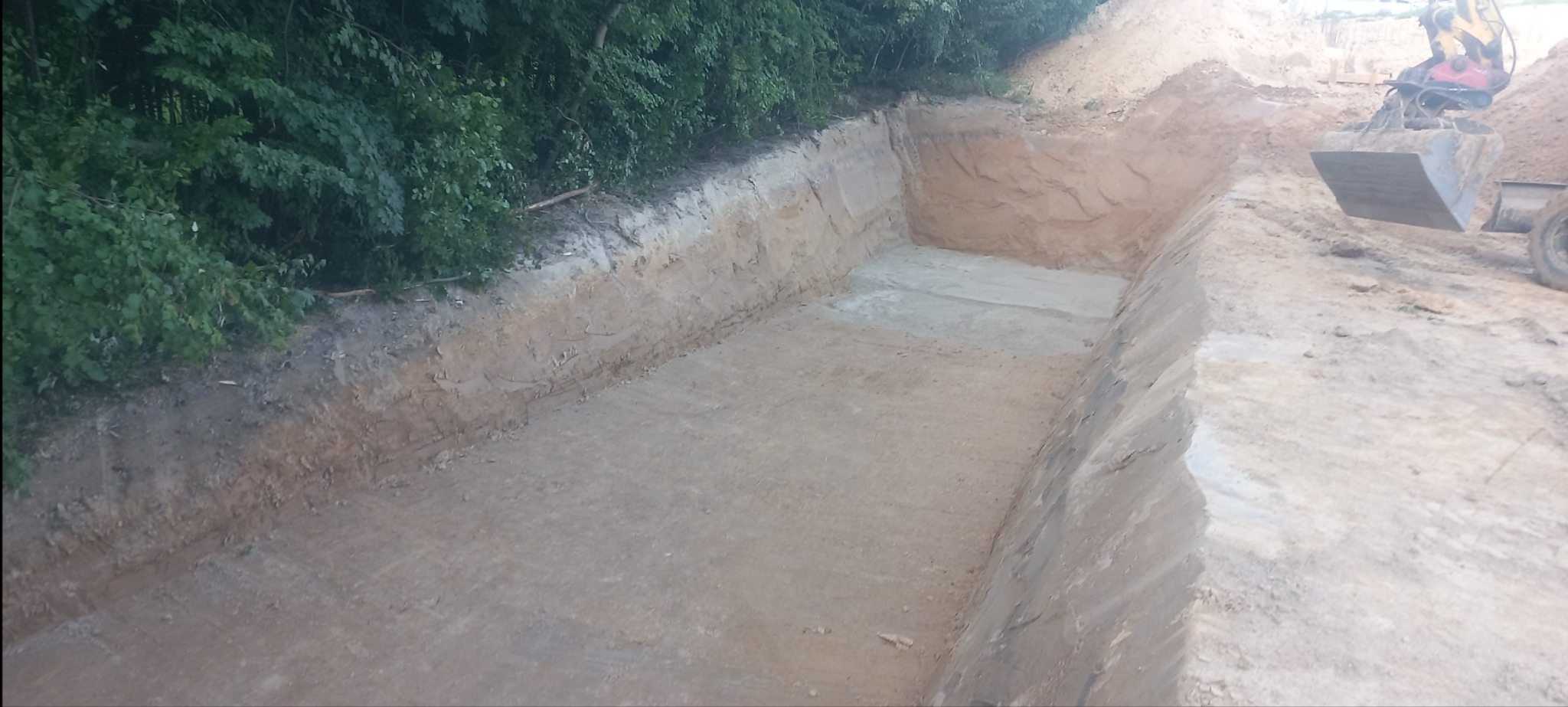 Prostokątny wykop w ziemi, z zielenią w tle i koparką w prawym górnym rogu, ukazujący etap prac ziemnych.