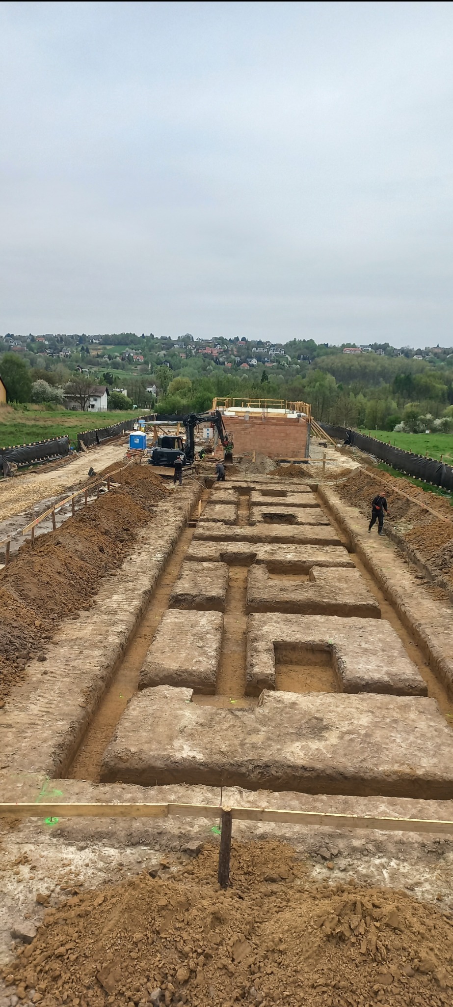 Wykop pod fundamenty domu jednorodzinnego z widocznym wykopem o skomplikowanym kształcie, deskowaniem i pracującą koparką w tle, na tle zielonych wzgórz.