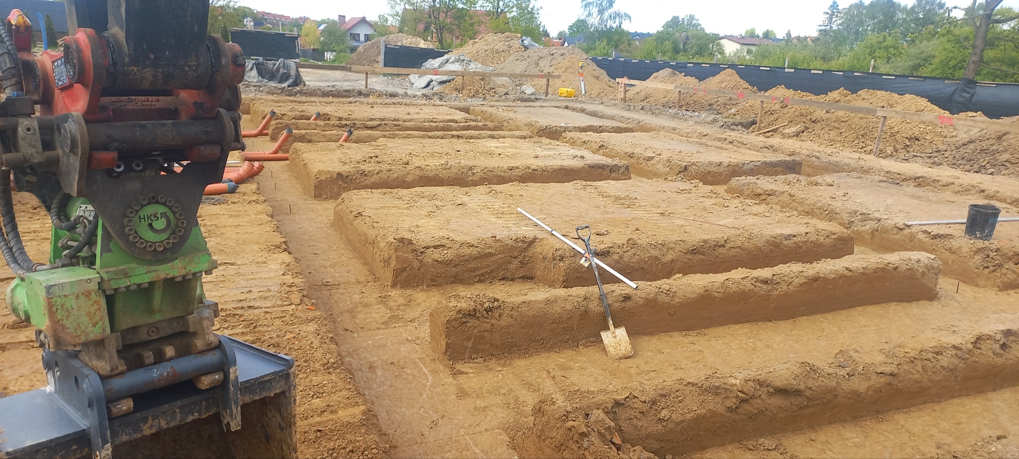Przygotowane wykopy pod fundamenty z widocznymi rurami drenażowymi, łopatą i łatą pomiarową, sprzęt budowlany w lewym rogu, widok na teren budowy.