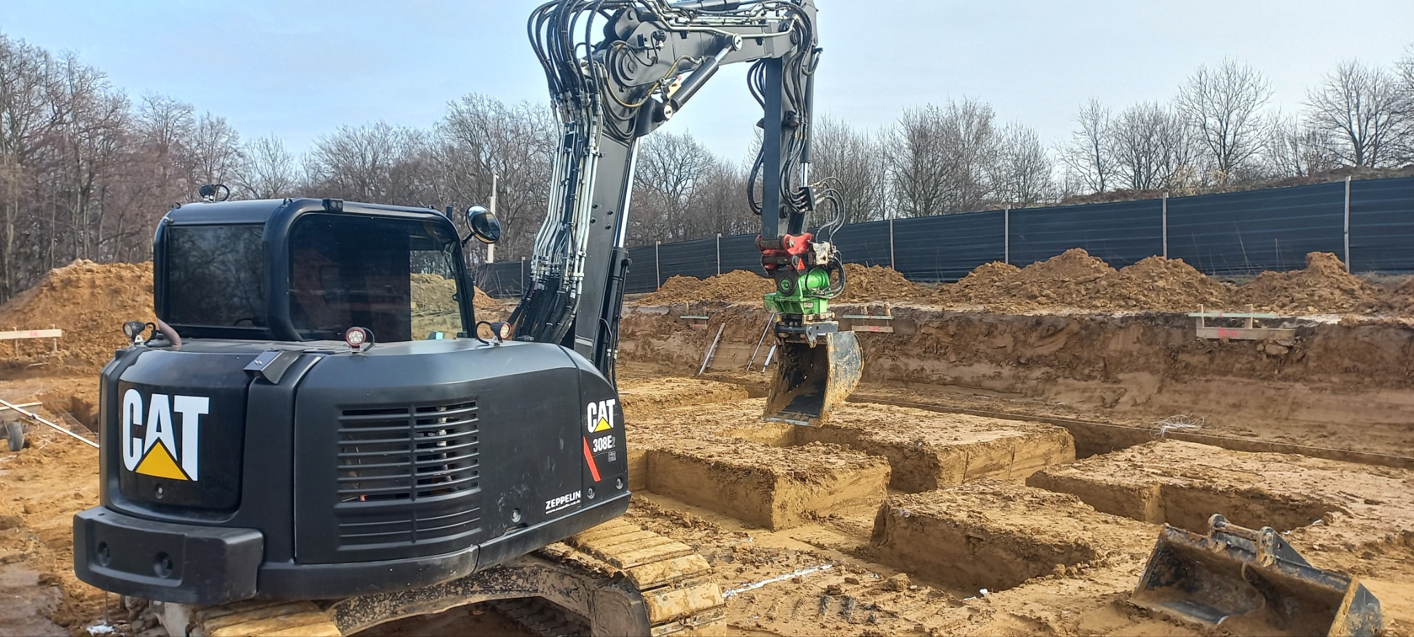 Czarna koparka CAT 308E2 CR z zielonym osprzętem kopiąca fundamenty pod budynek na placu budowy, widoczne wykopane bloki ziemi i zabezpieczenia wykopu.