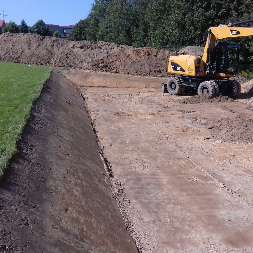 Żółta koparka CAT na brzegu wykopu, z pryzmą ziemi w tle, widok z boku na wyrobisko.