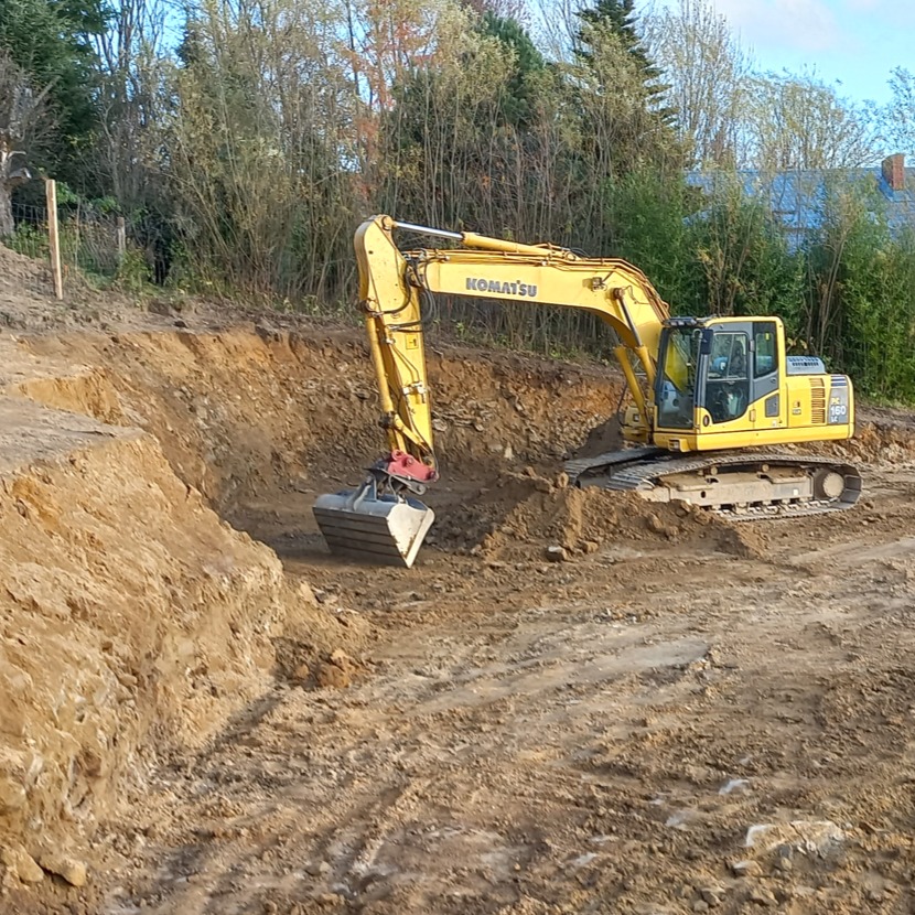 Żółta koparka Komatsu PC 160 pracuje przy wykopie w ziemi, w tle widoczne drzewa i budynek.