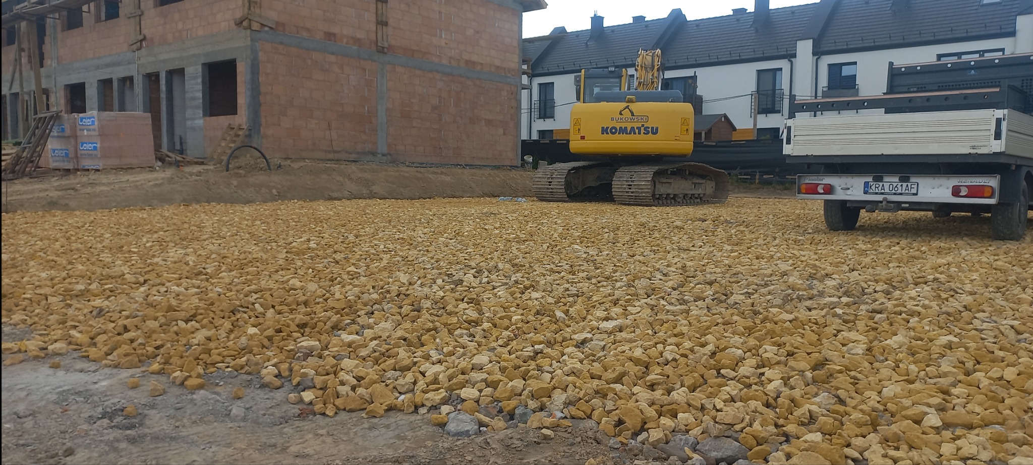 Żółty żwir równomiernie rozłożony na placu budowy z widoczną koparką Komatsu i ciężarówką Renault Master z tablicą rejestracyjną KRA 061AF, w tle budynek w trakcie budowy z cegły.