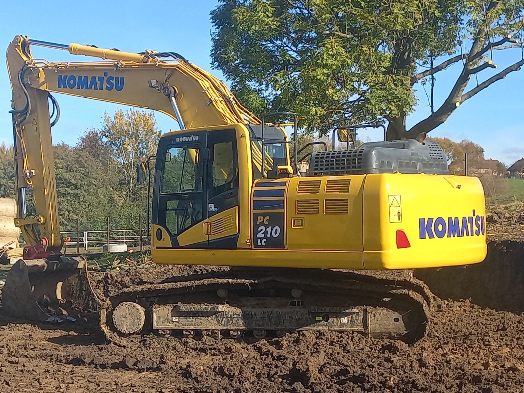 Żółta koparka Komatsu PC210 LC na gąsienicach stoi w wykopie, z drzewem w tle pod błękitnym niebem.