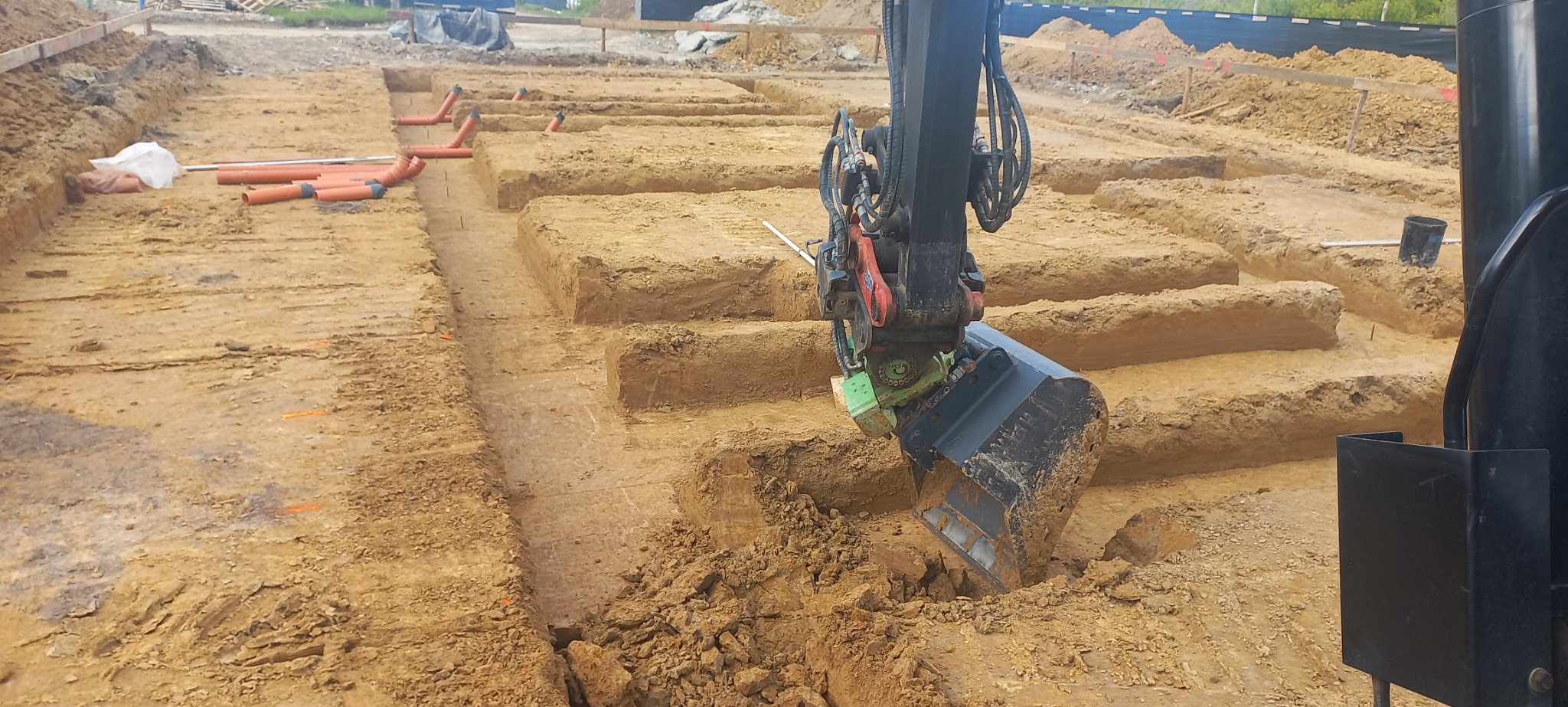 Mini koparka z czarną łyżką pogłębia wykop fundamentowy pod budynek, widoczne rury kanalizacyjne i ślady po wytyczeniu osi budynku.