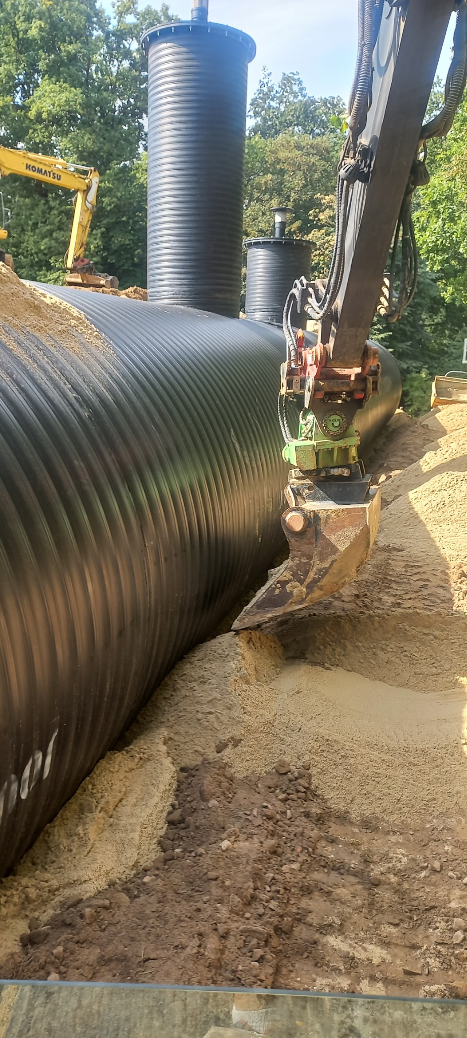 Ekskawator zasypuje piaskiem duży, czarny, karbowany zbiornik podziemny, widoczne pionowe rury wentylacyjne i inny sprzęt budowlany w tle.