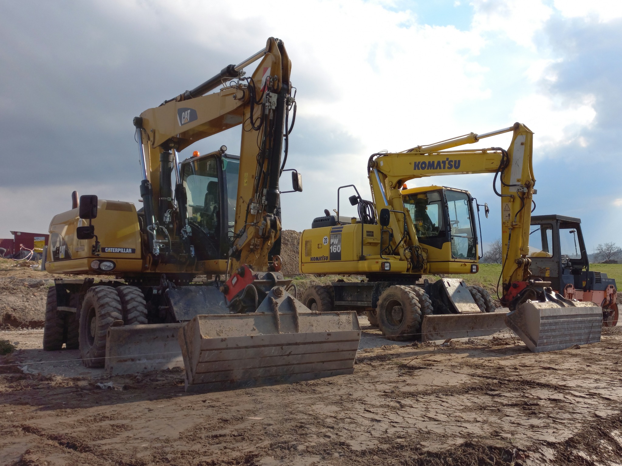 Dwie żółte koparki kołowe Caterpillar i Komatsu stoją na ubitej, gliniastej ziemi pod lekko pochmurnym niebem, widoczny walec w tle.