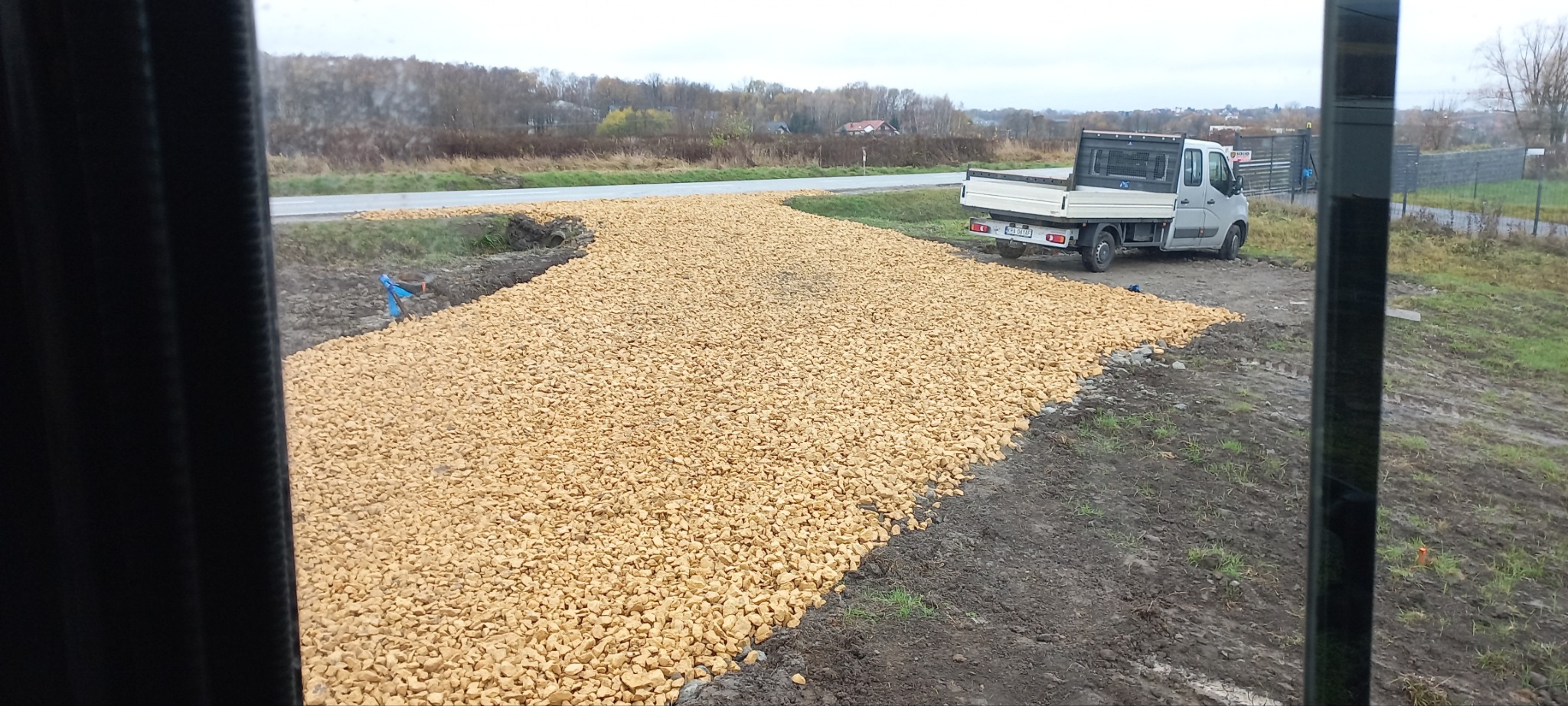 Żwirowa droga dojazdowa z zaparkowanym samochodem dostawczym na tle krajobrazu wiejskiego widziana przez okno.