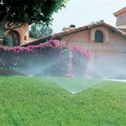 Zraszacze rotacyjne nawadniające trawnik przed domem z czerwonymi kwiatami i garażem w tle.