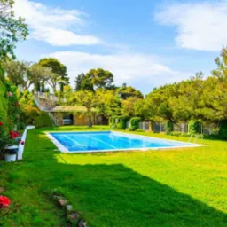 Prostokątny basen z błękitną wodą w ogrodzie z zielonym trawnikiem, otoczony drzewami i roślinnością, z widocznymi schodkami wejściowymi i filtrem piaskowym w tle.